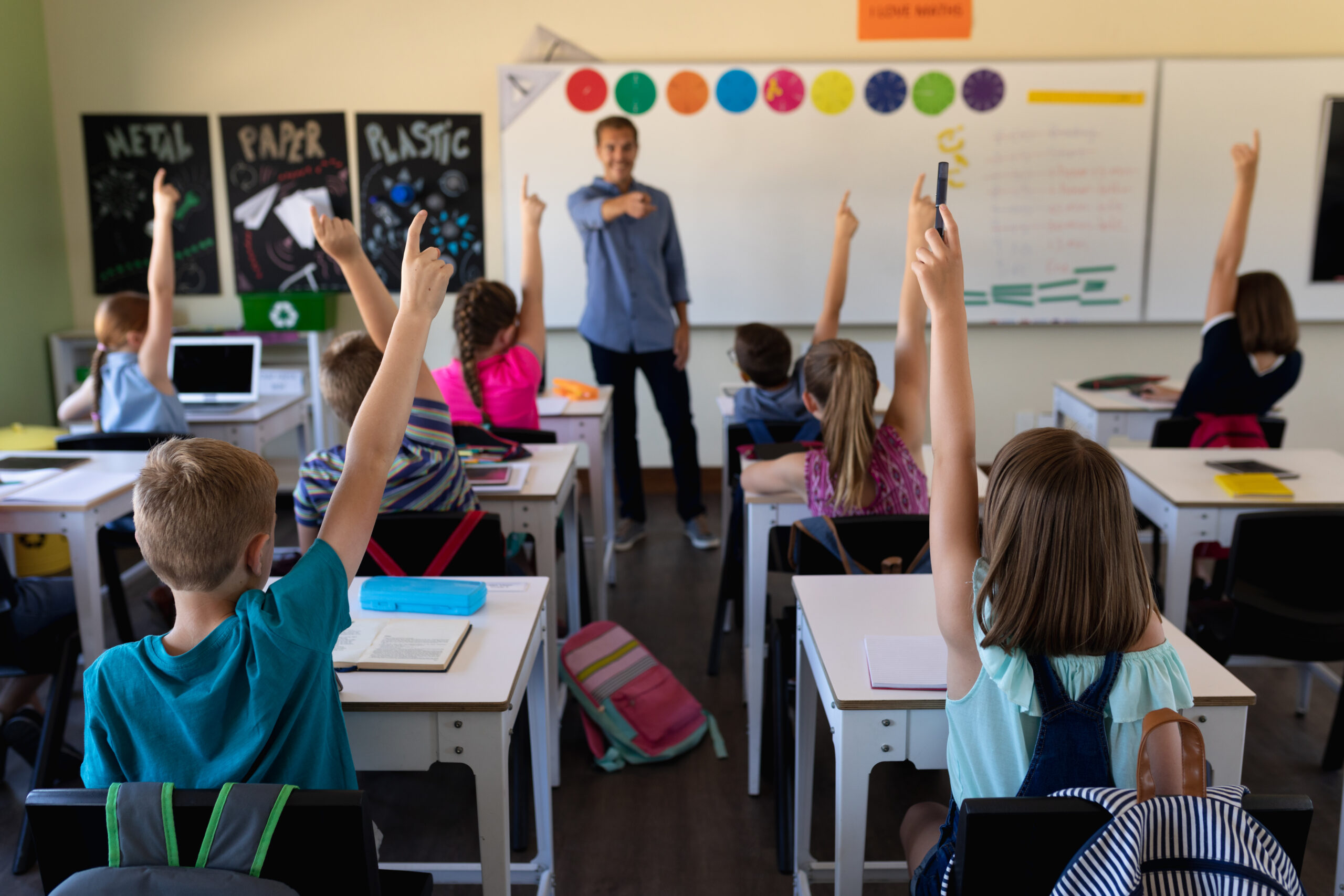 Front view of a Caucasian male school teacher standing in front of a diverse group of school children sitting at desks and raising their hands to answer a question during a lesson in an elementary school classroom, the teacher pointing to one of them to an